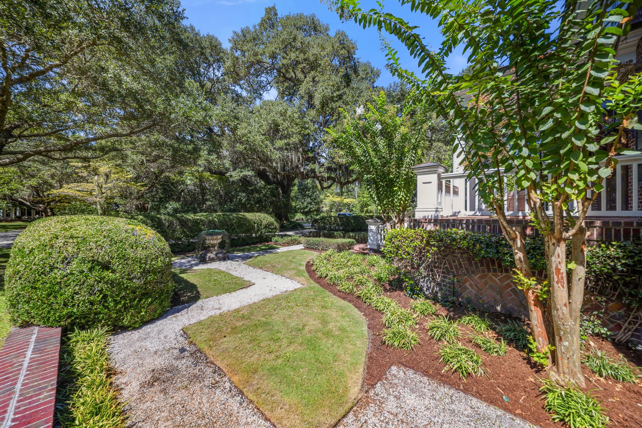 Garden path curving beneath large oak trees, with rounded clipped boxwood shrubs along the edge.