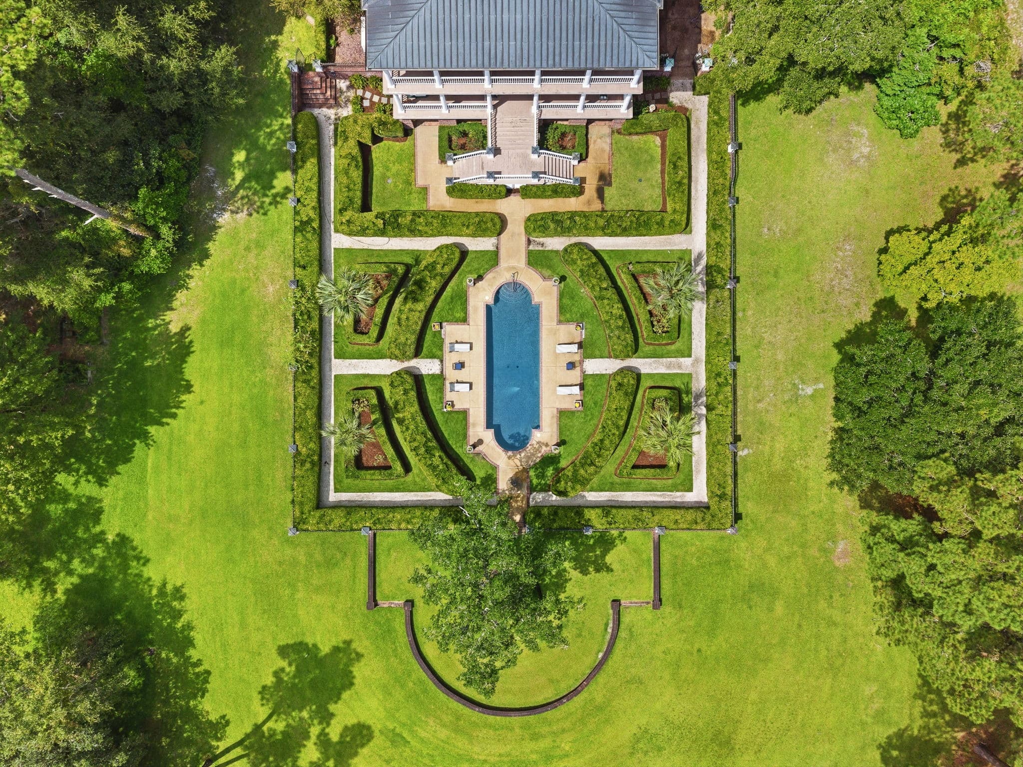 Top-down aerial view of a rectangular walled garden with an oval pool and clipped hedge beds, house roof at the edge of frame.