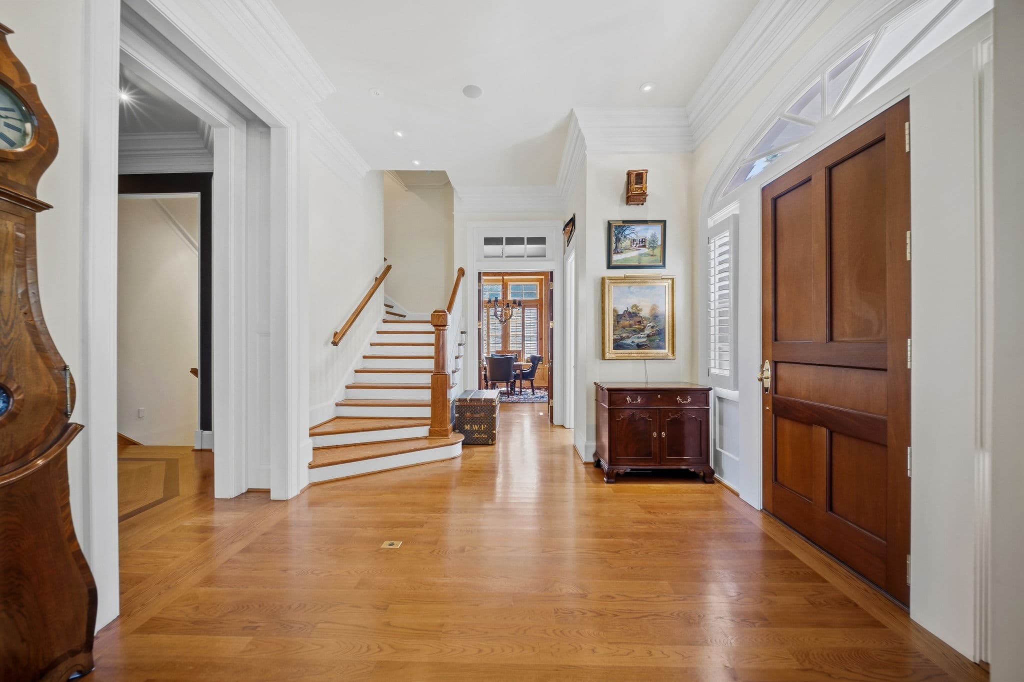 Hallway view showing a staircase, framed artwork on the wall, and a chandelier visible in a room at the end of the hall.