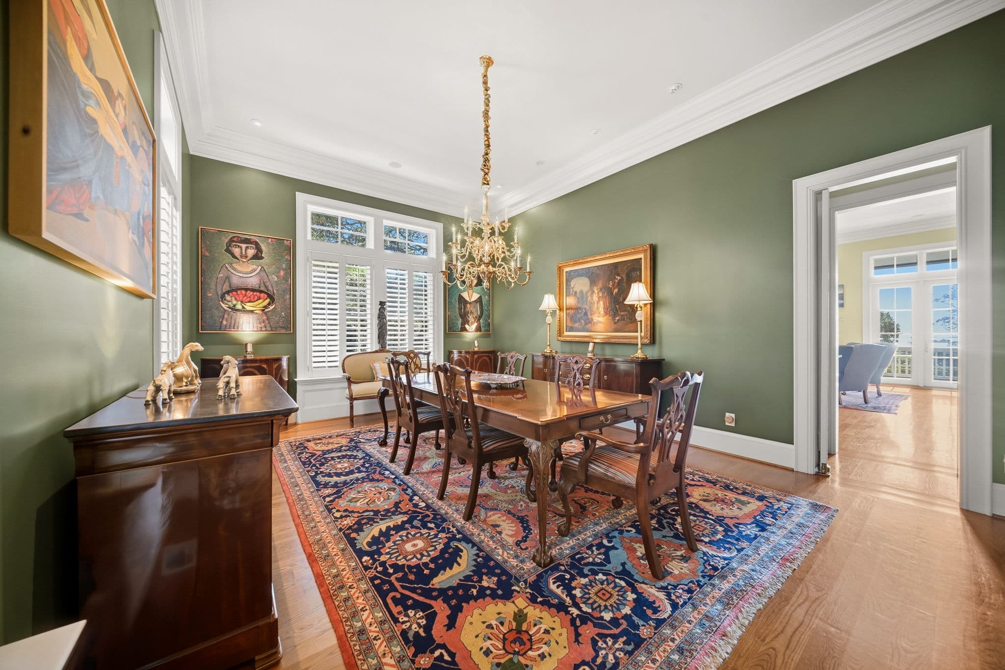 Dining room table and upholstered chairs on a patterned rug, with framed artwork on green walls.