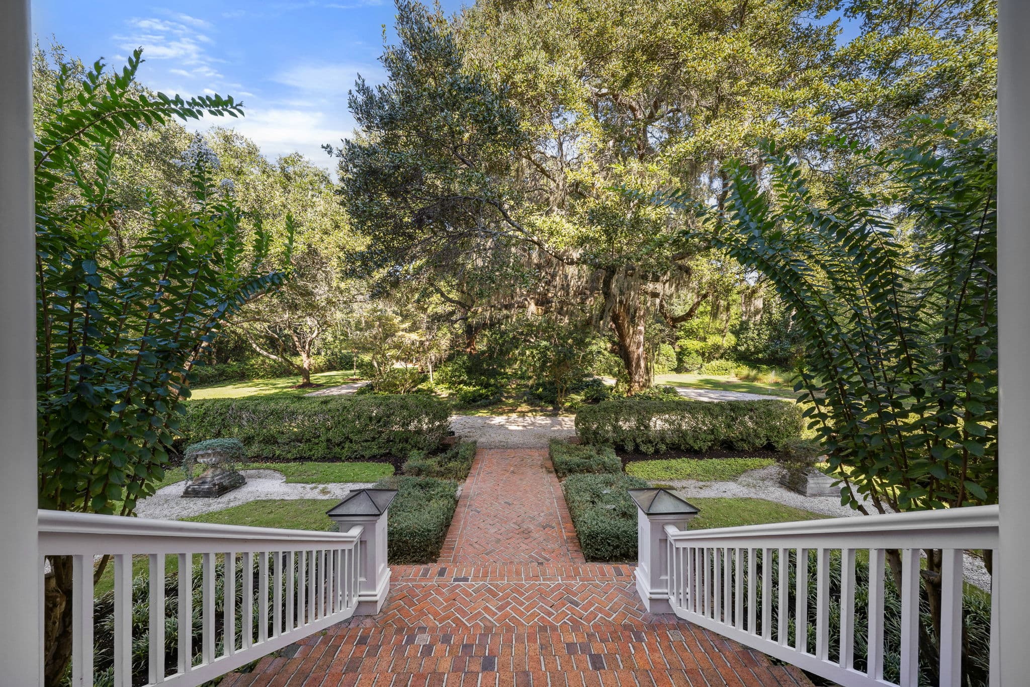 View from the porch through white railings and ferns, overlooking a brick herringbone path and boxwood garden.