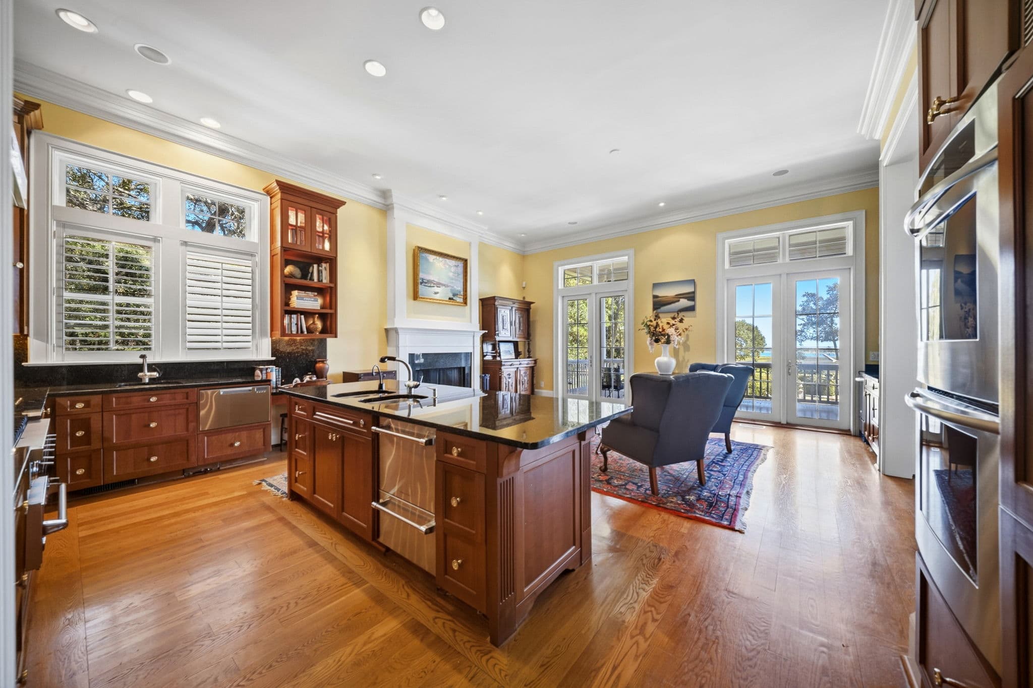 Sitting area beside the kitchen with wingback chairs, a fireplace, and French doors showing a water view in the distance.