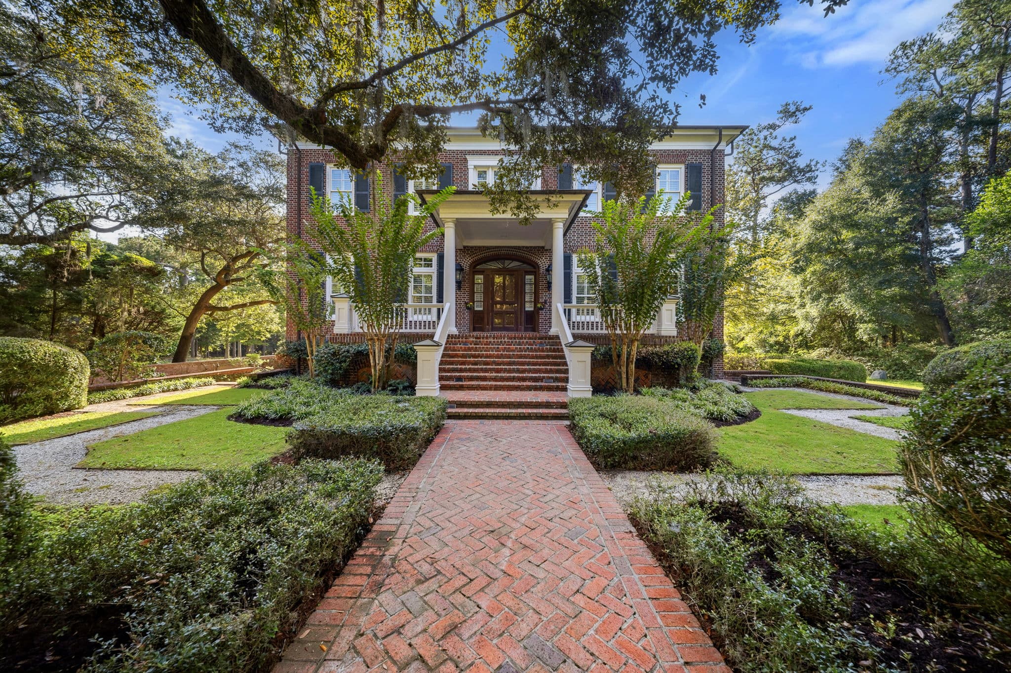 Front exterior of a two story brick home with white portico columns and a brick herringbone front walkway, framed by an oak tree.