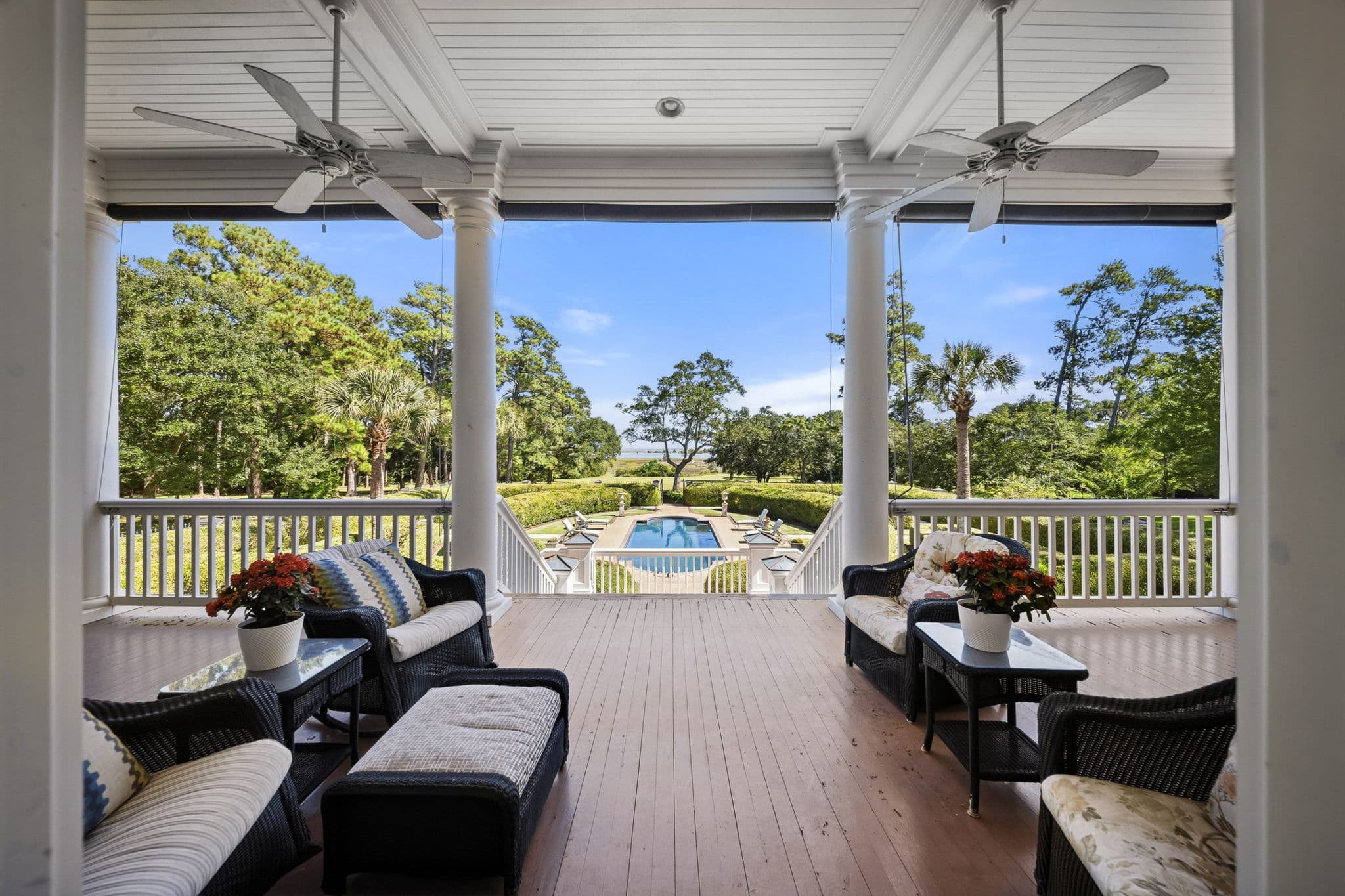 Covered porch with ceiling fans and wicker furniture overlooking a rectangular pool and gardens.
