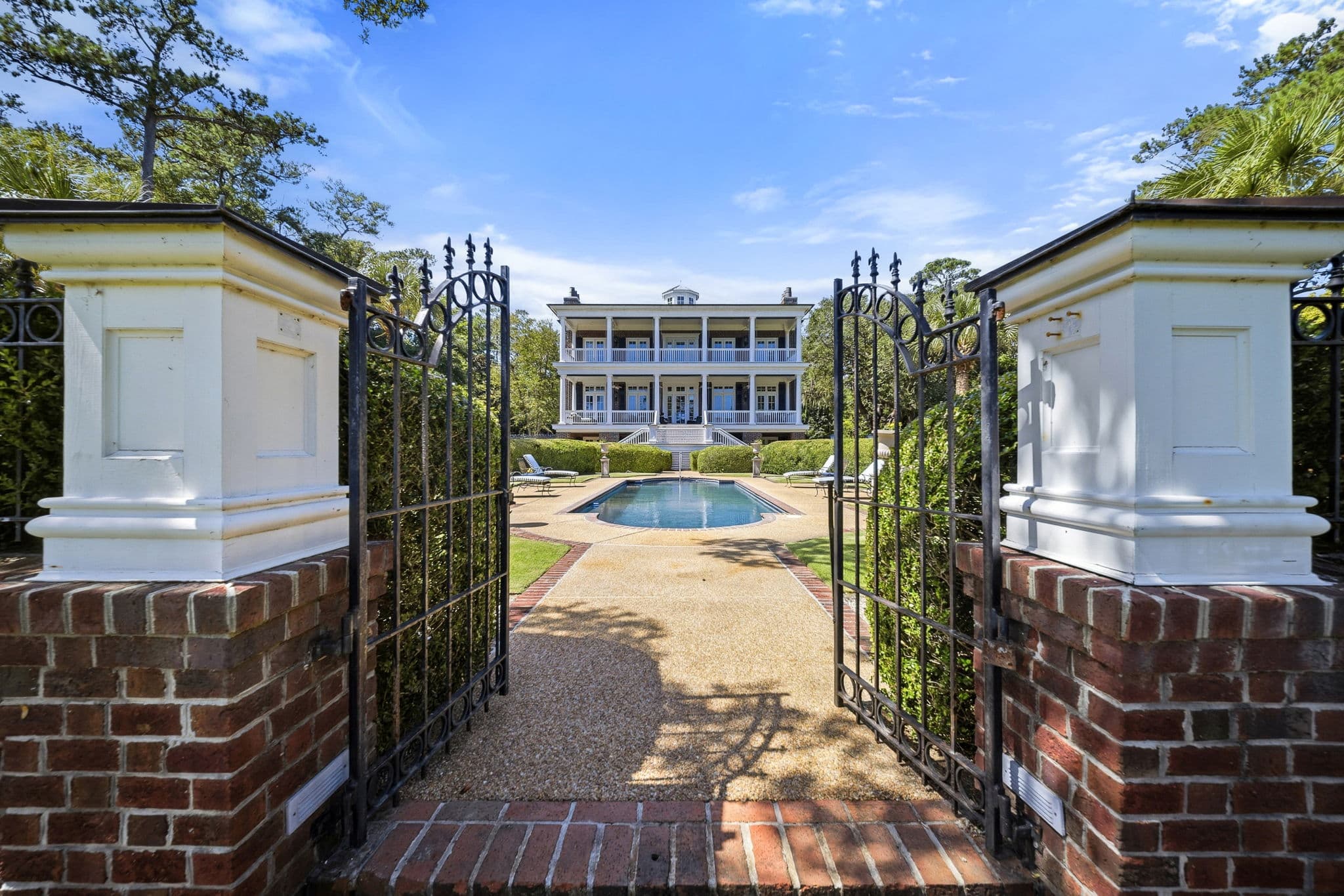 Three-story house with columned porches seen through an open garden gate, with a pool in between.