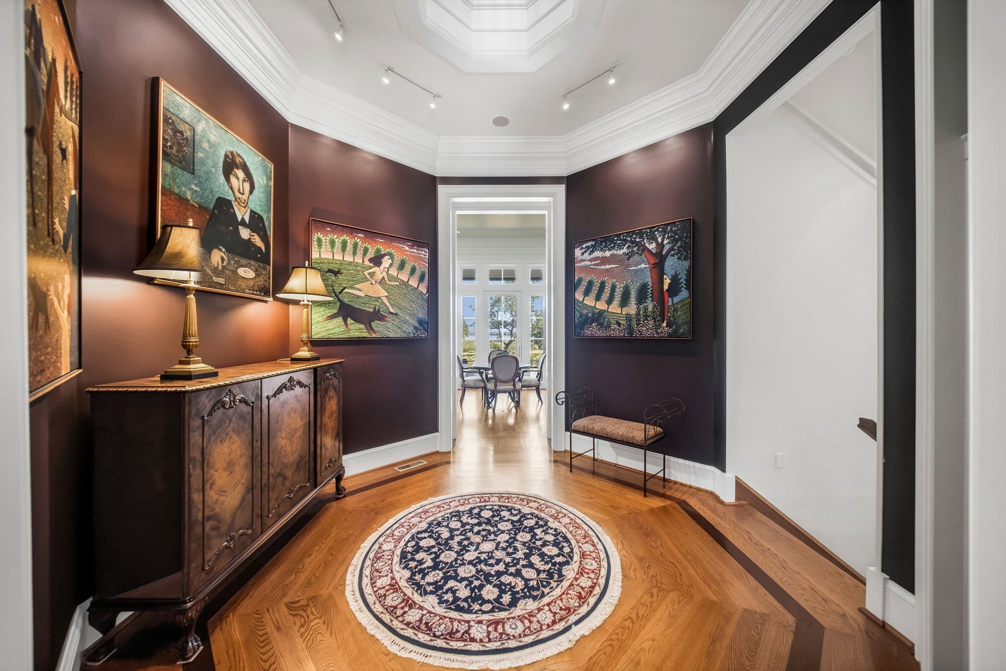 Octagonal hallway with a ceiling skylight, plum walls, framed paintings, and a wood sideboard with table lamps.