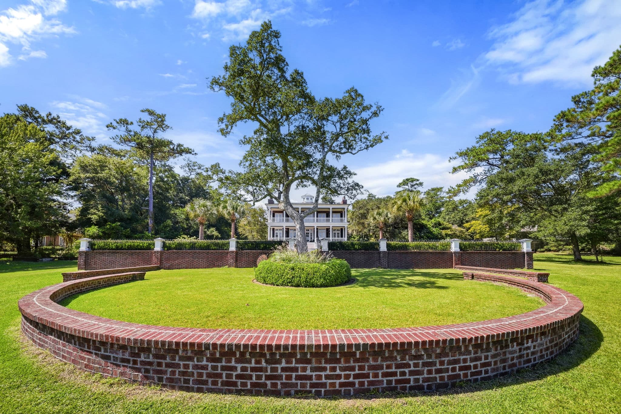 Close view of a curved brick wall and mature oak tree with the house and iron fence visible behind.