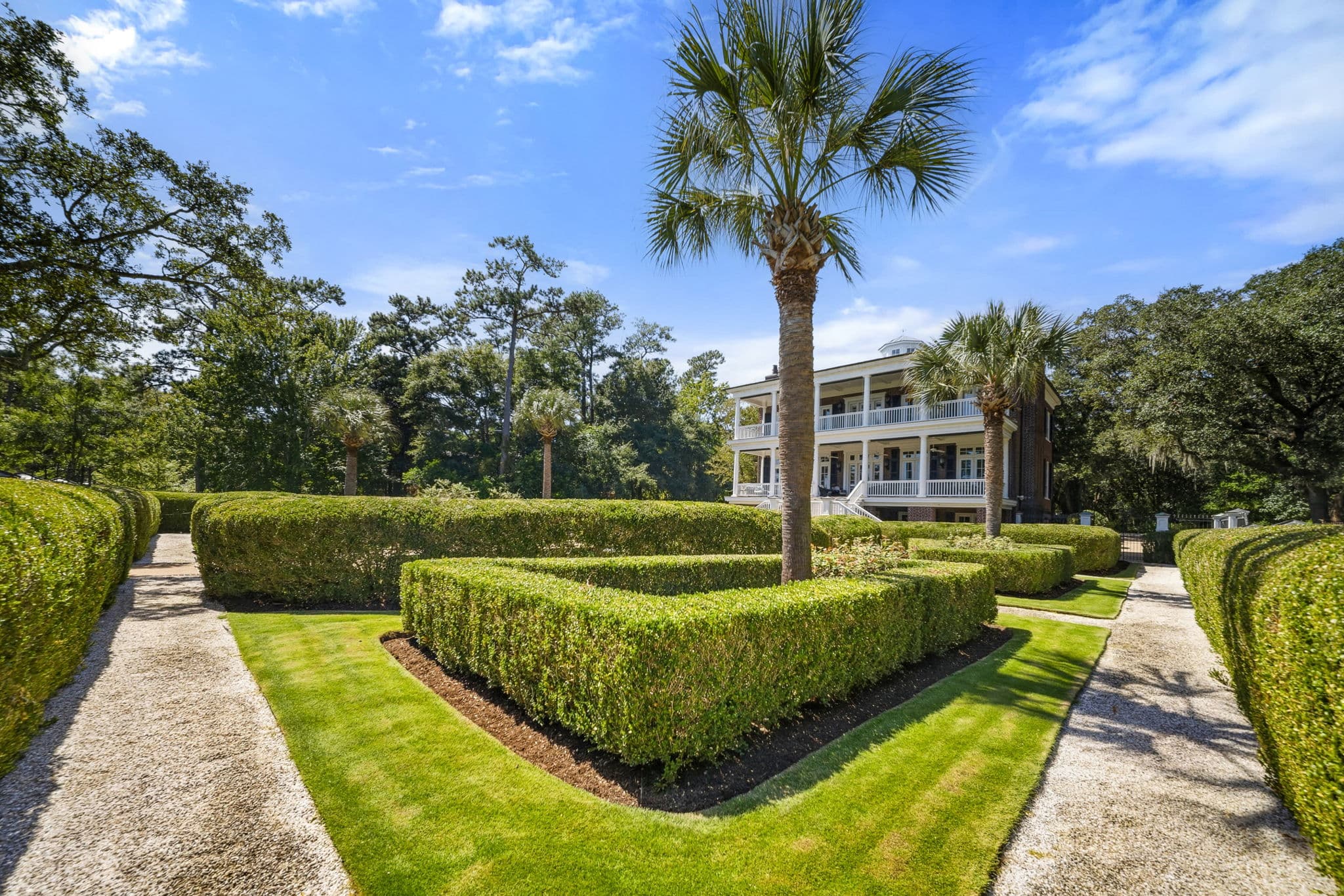 Garden with clipped hedge beds, a central palm tree, and gravel paths leading toward a house with tiered porches.