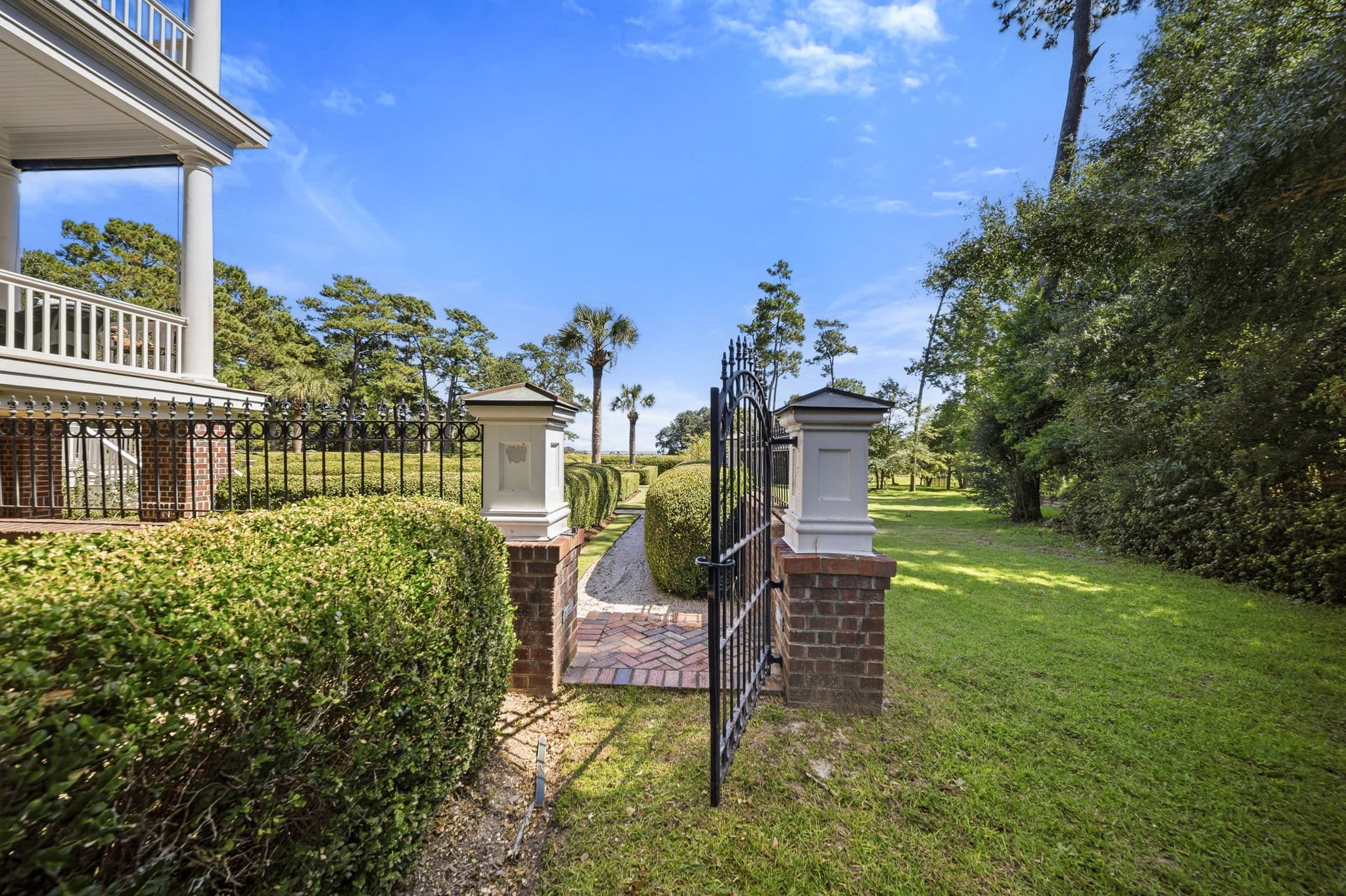 Open wrought-iron garden gate flanked by brick posts, with a hedge-lined path leading toward the marsh view.