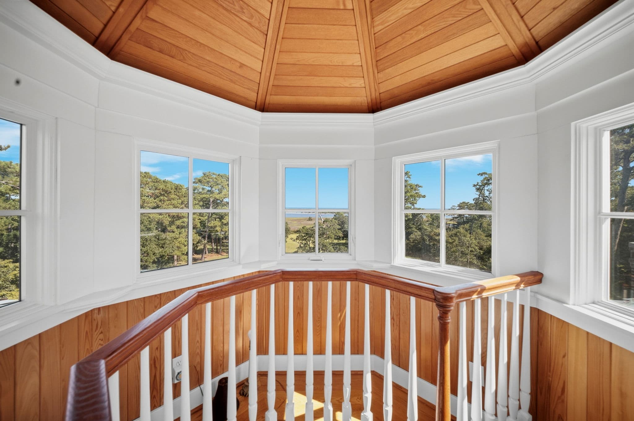 Octagonal room at the top of the house with windows on all sides and a wood plank ceiling, overlooking trees and water.