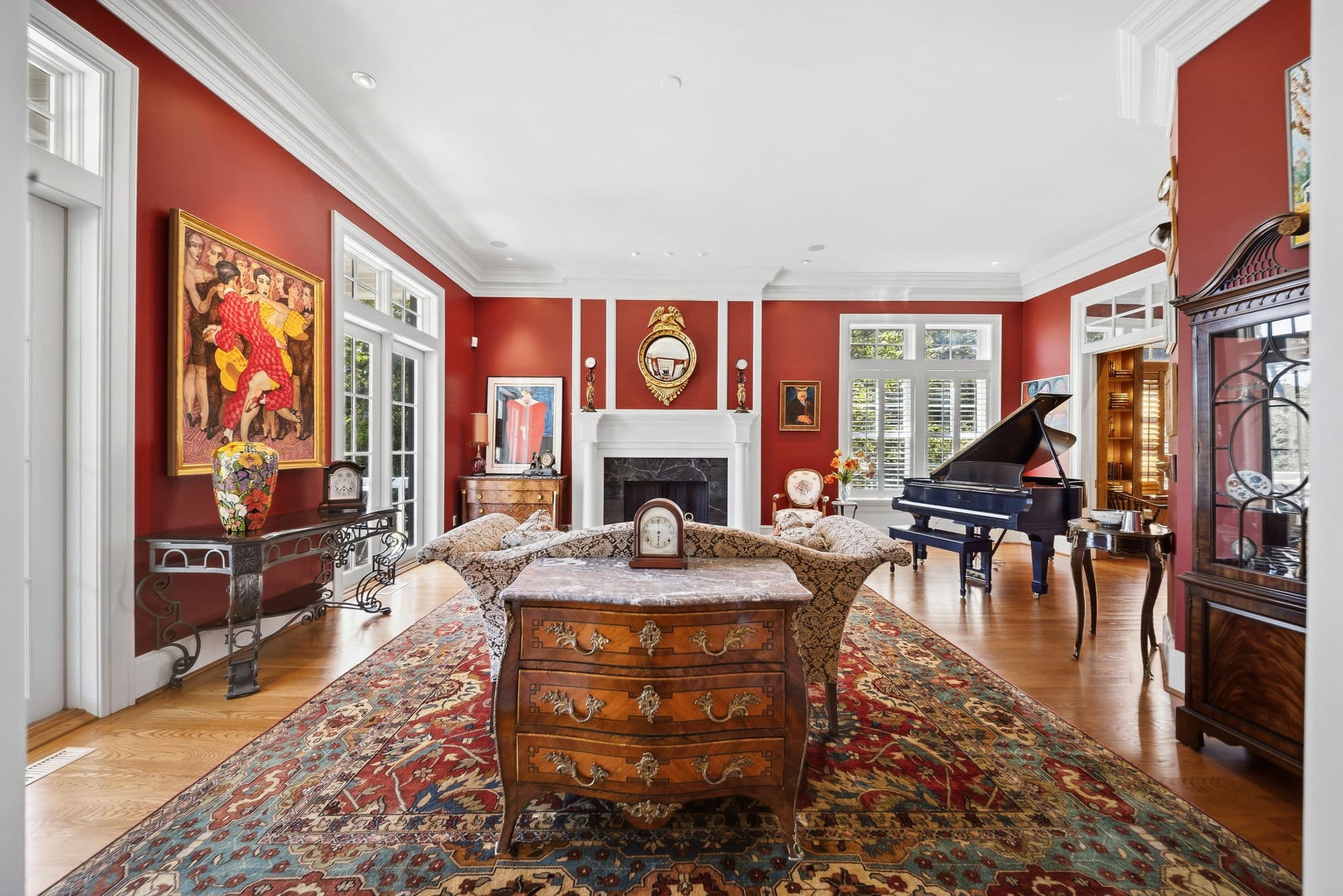 Red living room with a marble-top chest and floral vase in the foreground, a gilt mirror over the fireplace, and a piano in the adjoining room.