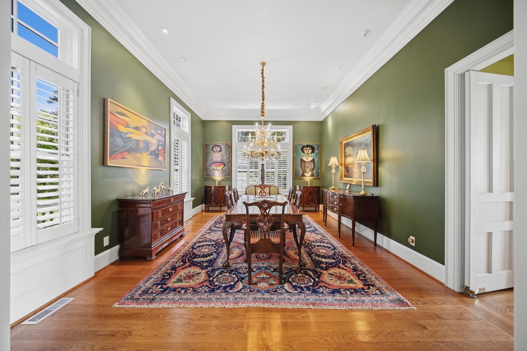 Dining room with brass chandelier, long wood table and chairs, and shuttered windows.