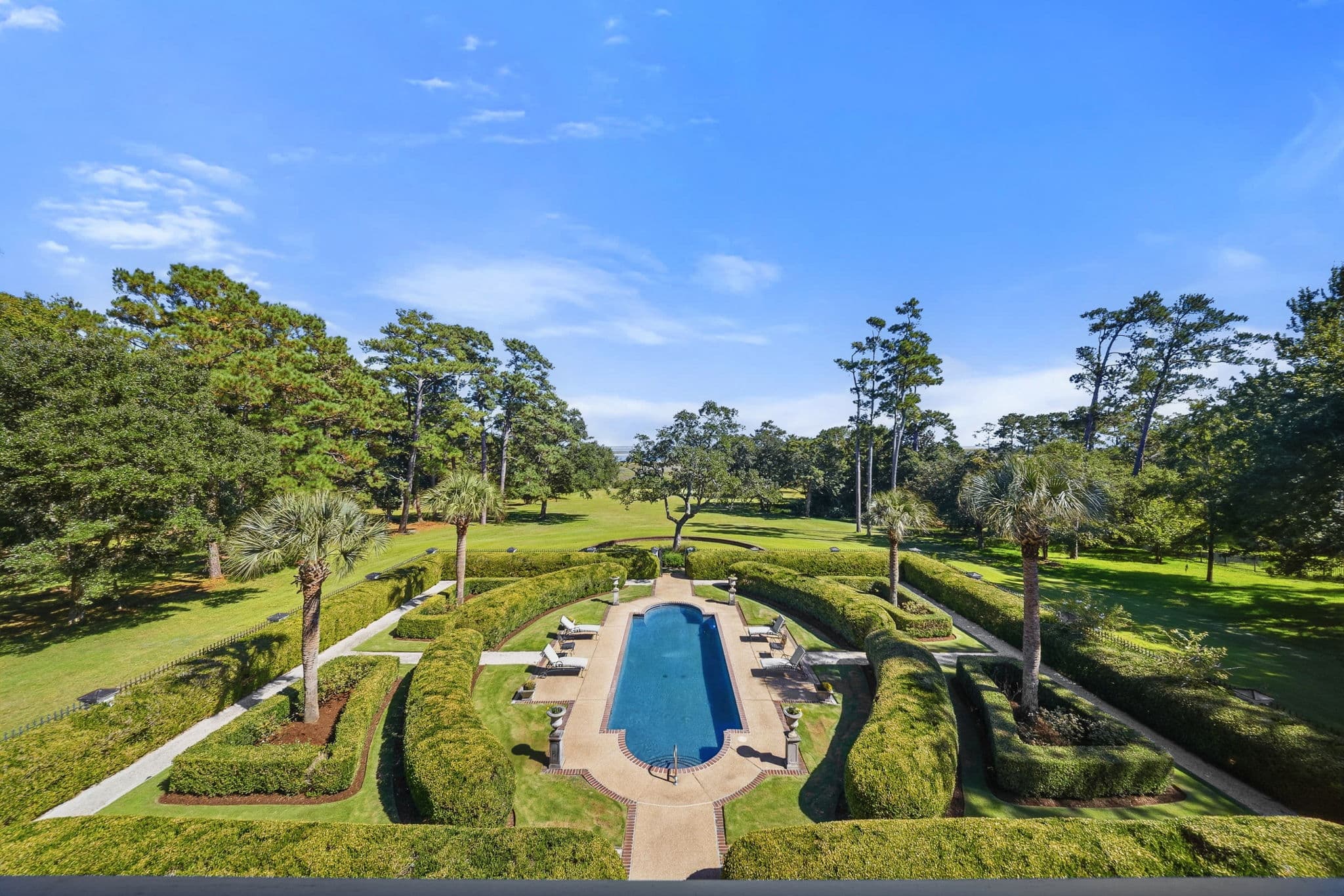 Aerial view of the pool and clipped hedge garden with pine trees and a distant shoreline.