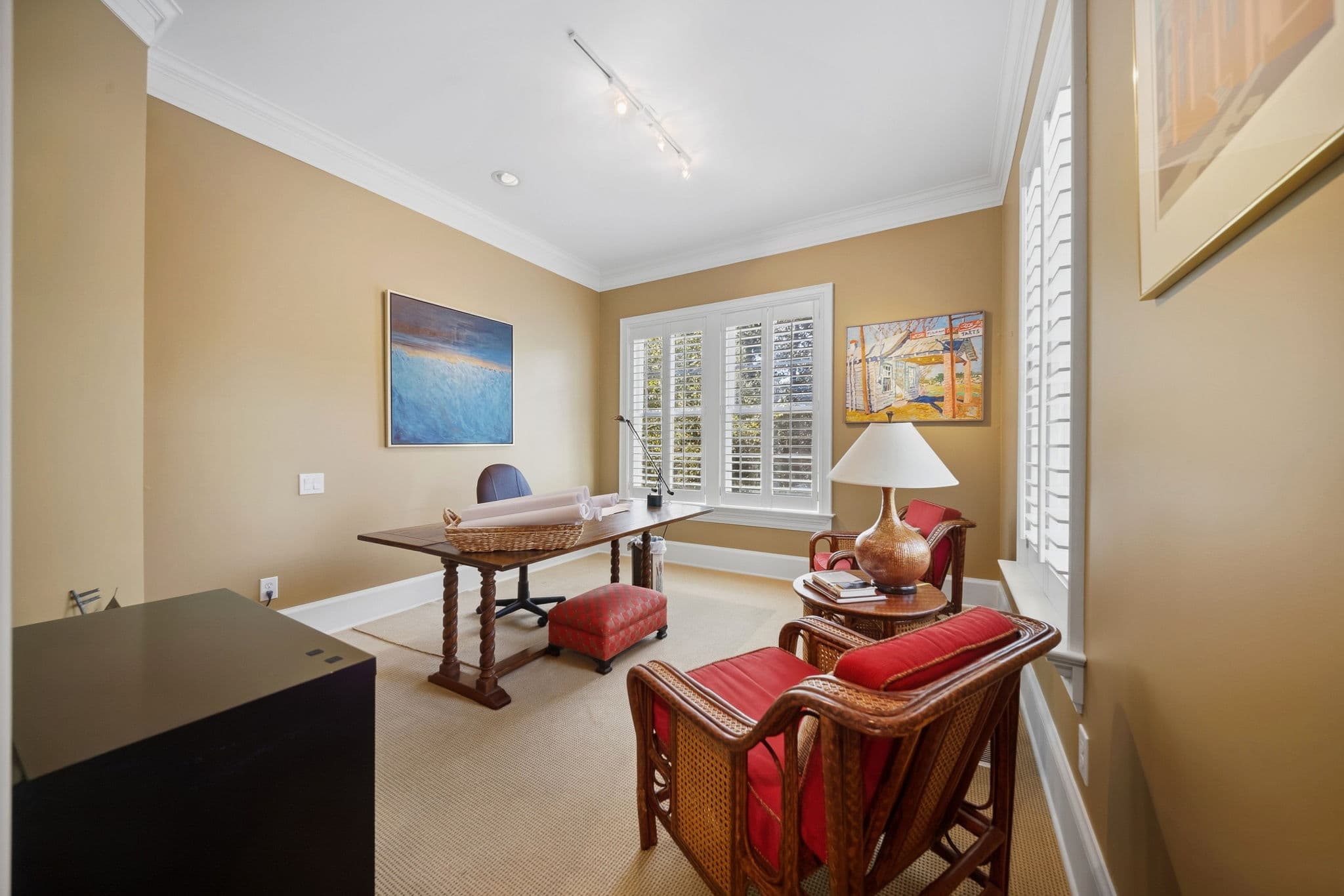 Home office with a wood trestle desk, rattan armchairs with red cushions, and framed artwork.